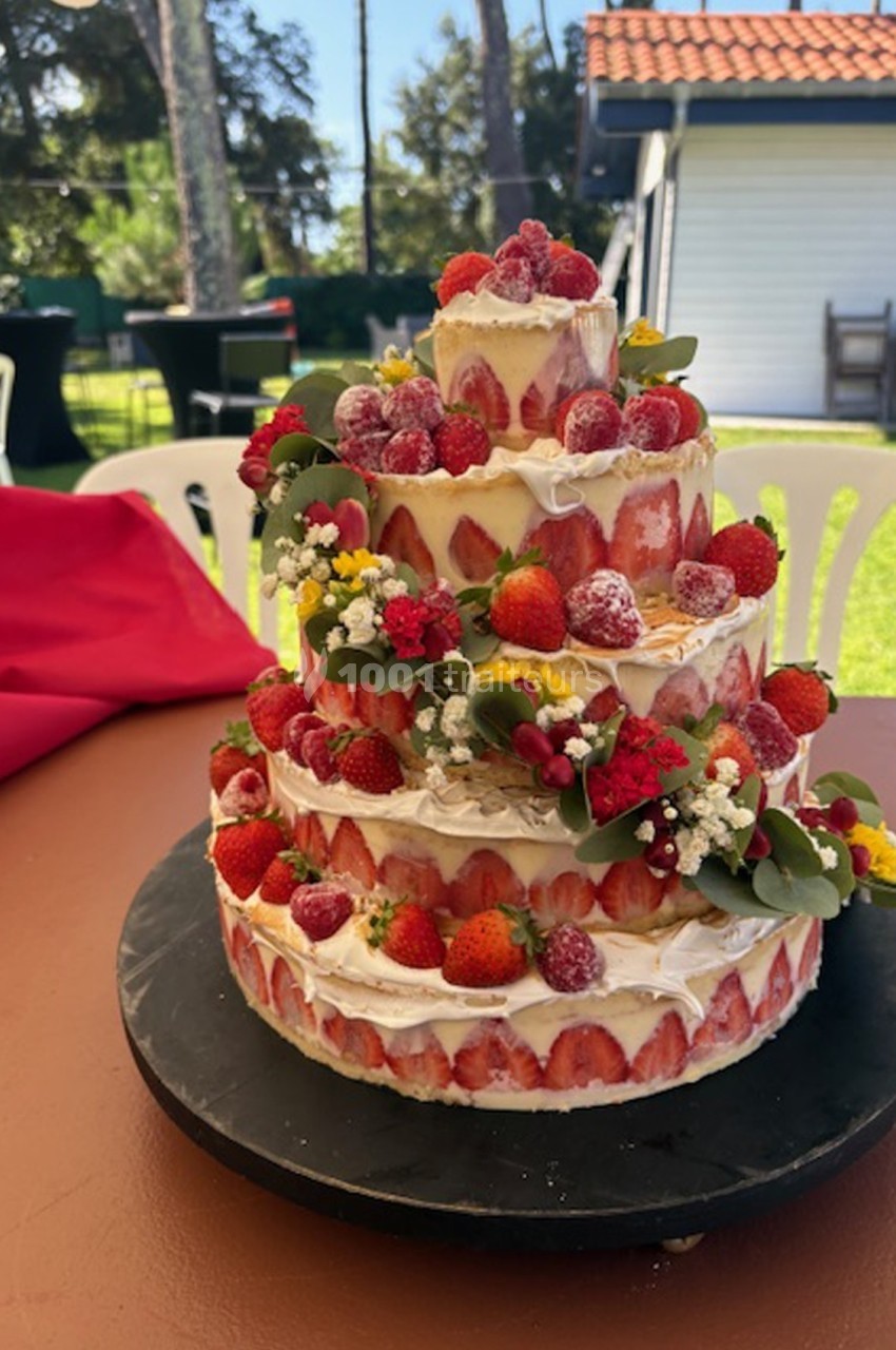 Gâteau à étages décoré de fraises, framboises et fleurs, présenté sur une table en extérieur par temps ensoleillé.