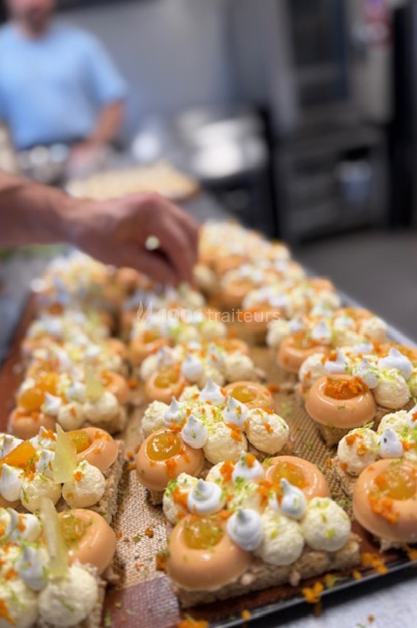 Des pâtisseries garnies de crème et de fruits sont disposées sur un plateau, une main ajoute des décorations.