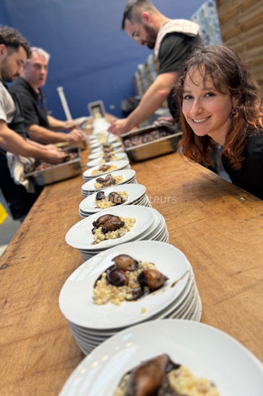 Des assiettes alignées contenant un plat à base de céréales et légumes, avec des cuisiniers en arrière-plan.