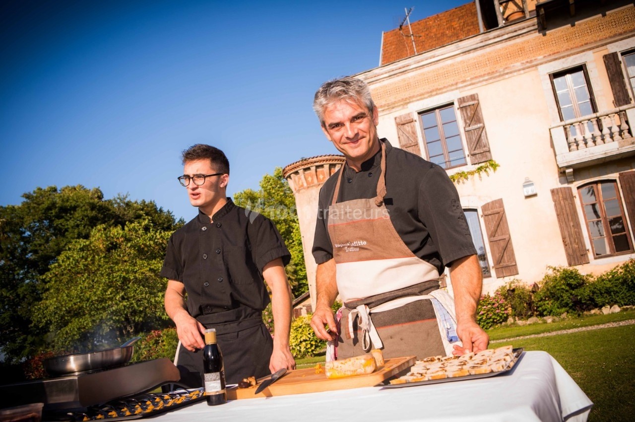 Deux chefs cuisinent en extérieur devant une grande maison, avec des ingrédients disposés sur une table.