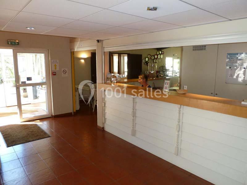 Accueil d'un établissement avec comptoir en bois, employée à son poste, porte vitrée et décoration sobre en arrière-plan.