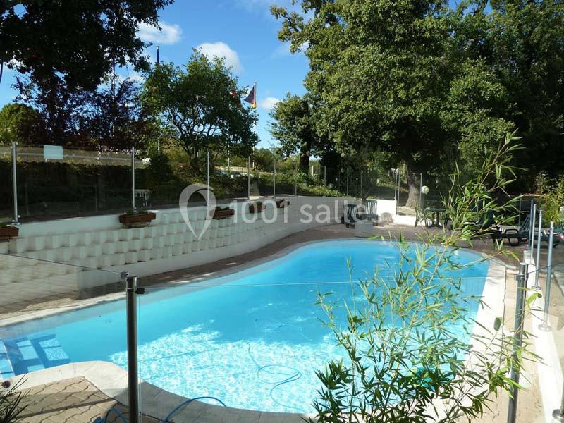 Piscine extérieure entourée de végétation, avec des chaises longues et une clôture en verre.