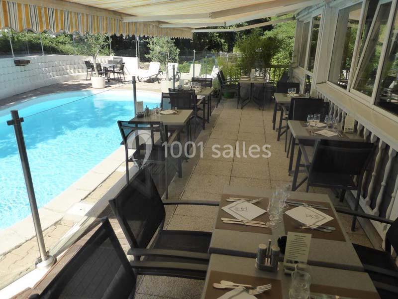 Terrasse d'un restaurant avec tables dressées, située près d'une piscine entourée de verdure et protégée par un store.