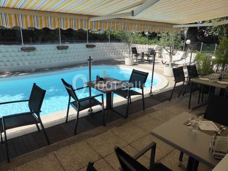 Terrasse ombragée avec tables et chaises donnant sur une piscine extérieure entourée de végétation et de transats.