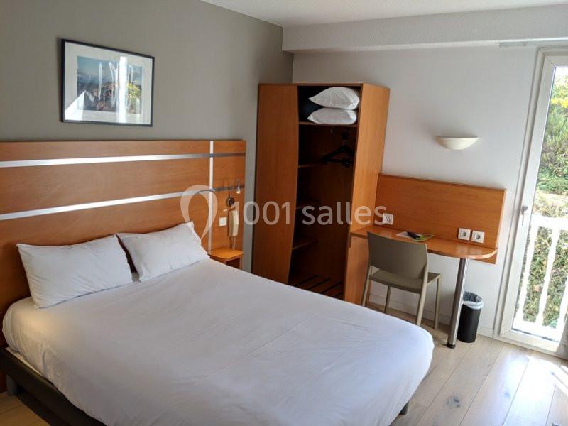 Chambre d'hôtel avec lit double, bureau, armoire ouverte, et lumière naturelle provenant d'une porte-fenêtre.