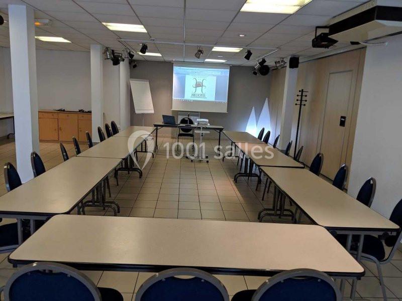 Salle de réunion en configuration en U avec tables, chaises, vidéoprojecteur et écran allumé.