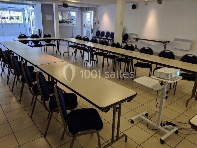 Salle de réunion lumineuse avec des tables disposées en U, des chaises alignées et un projecteur sur un support mobile.