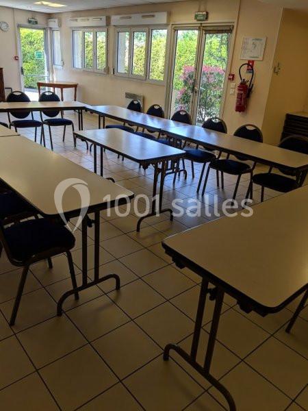Salle lumineuse avec tables rectangulaires et chaises disposées en U, fenêtres donnant sur un jardin.