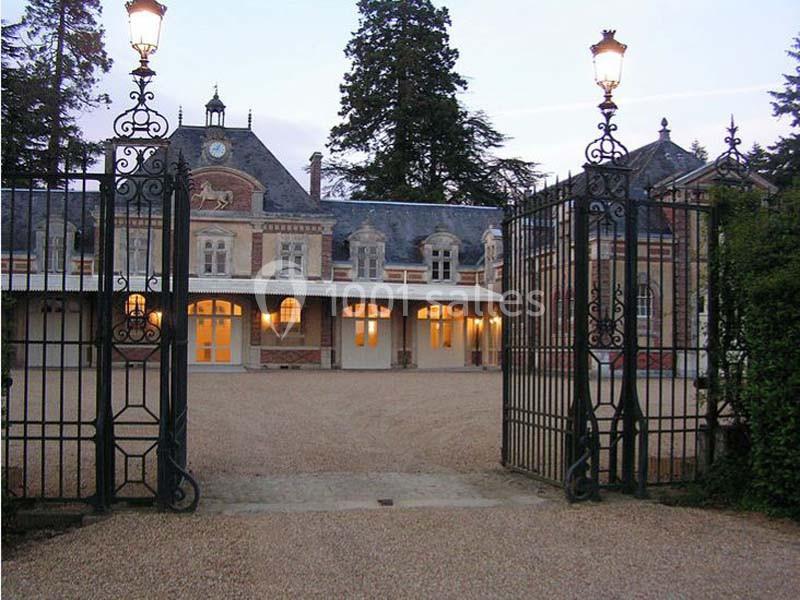 Entrée d'un domaine avec portail en fer forgé ouvert, donnant sur une cour gravillonnée et un bâtiment en arrière-plan.
