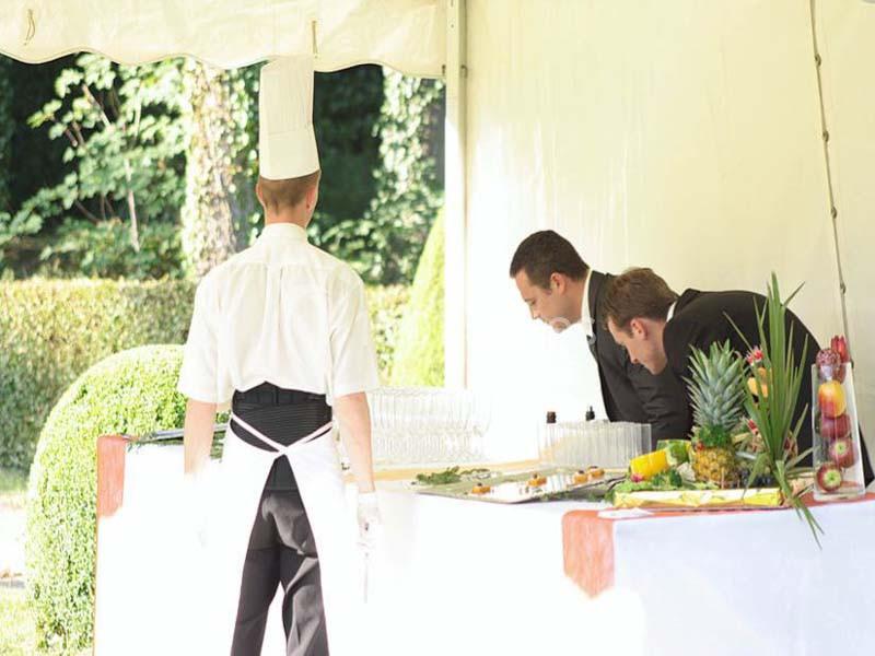 Un chef et deux hommes en costume préparent une table de buffet en extérieur sous une tente blanche.