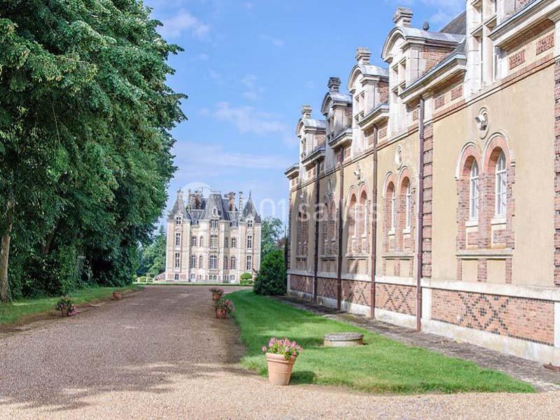 Allée bordée de pelouse menant à un château en pierre, avec des bâtiments annexes à droite et des arbres à gauche.