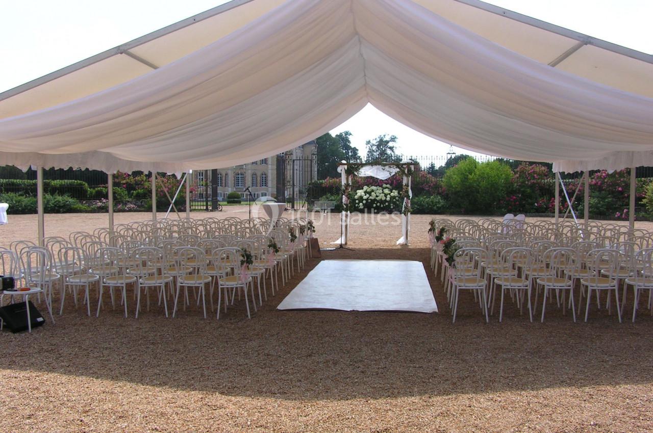 Tente blanche installée en extérieur avec des rangées de chaises et une arche décorée pour une cérémonie.