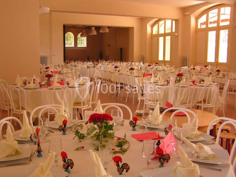 Salle de réception lumineuse avec tables rondes dressées, nappes blanches, serviettes pliées et décorations florales rouges.
