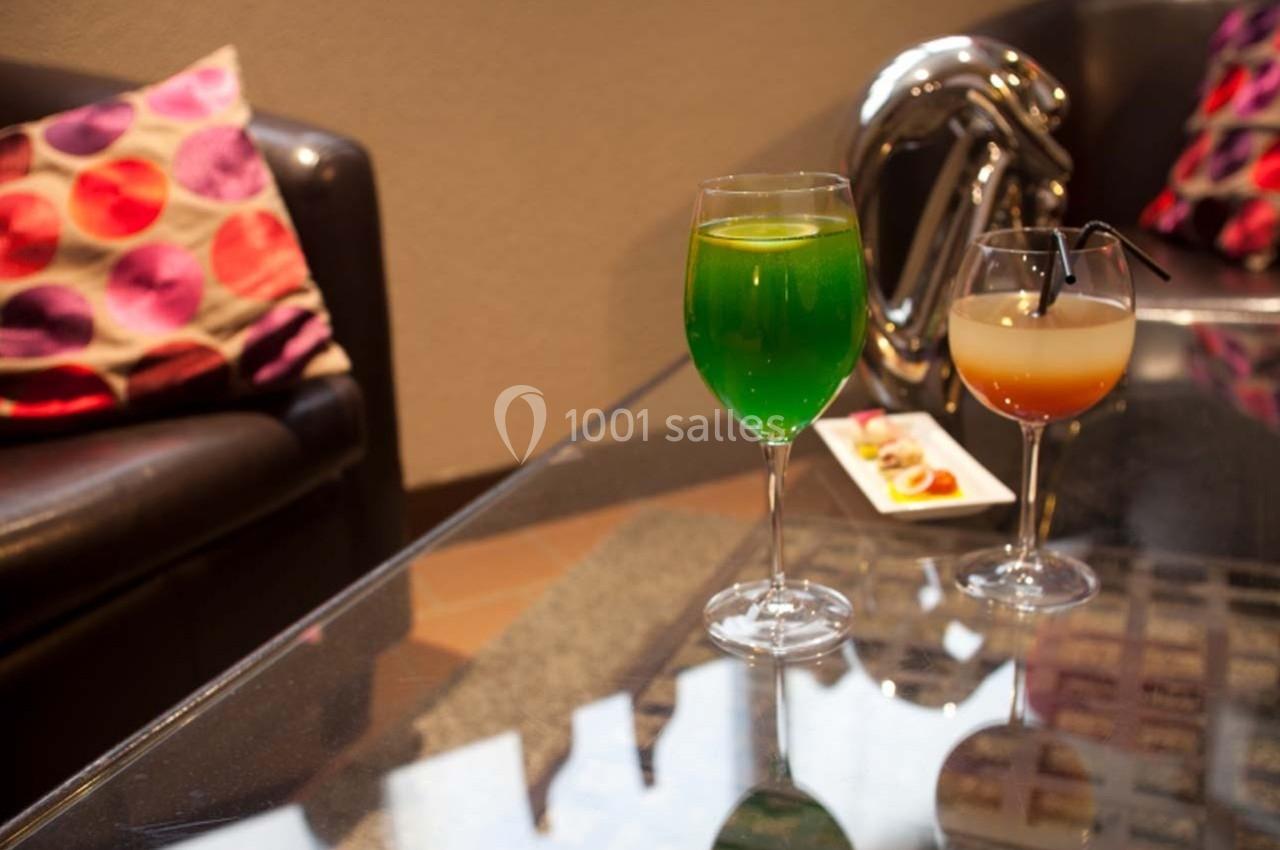 Deux verres de cocktails colorés posés sur une table en verre, avec un canapé et des coussins en arrière-plan.