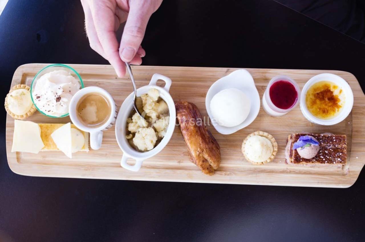 Plateau en bois avec divers desserts, café, fromage, et condiments, une main tenant une cuillère au centre.