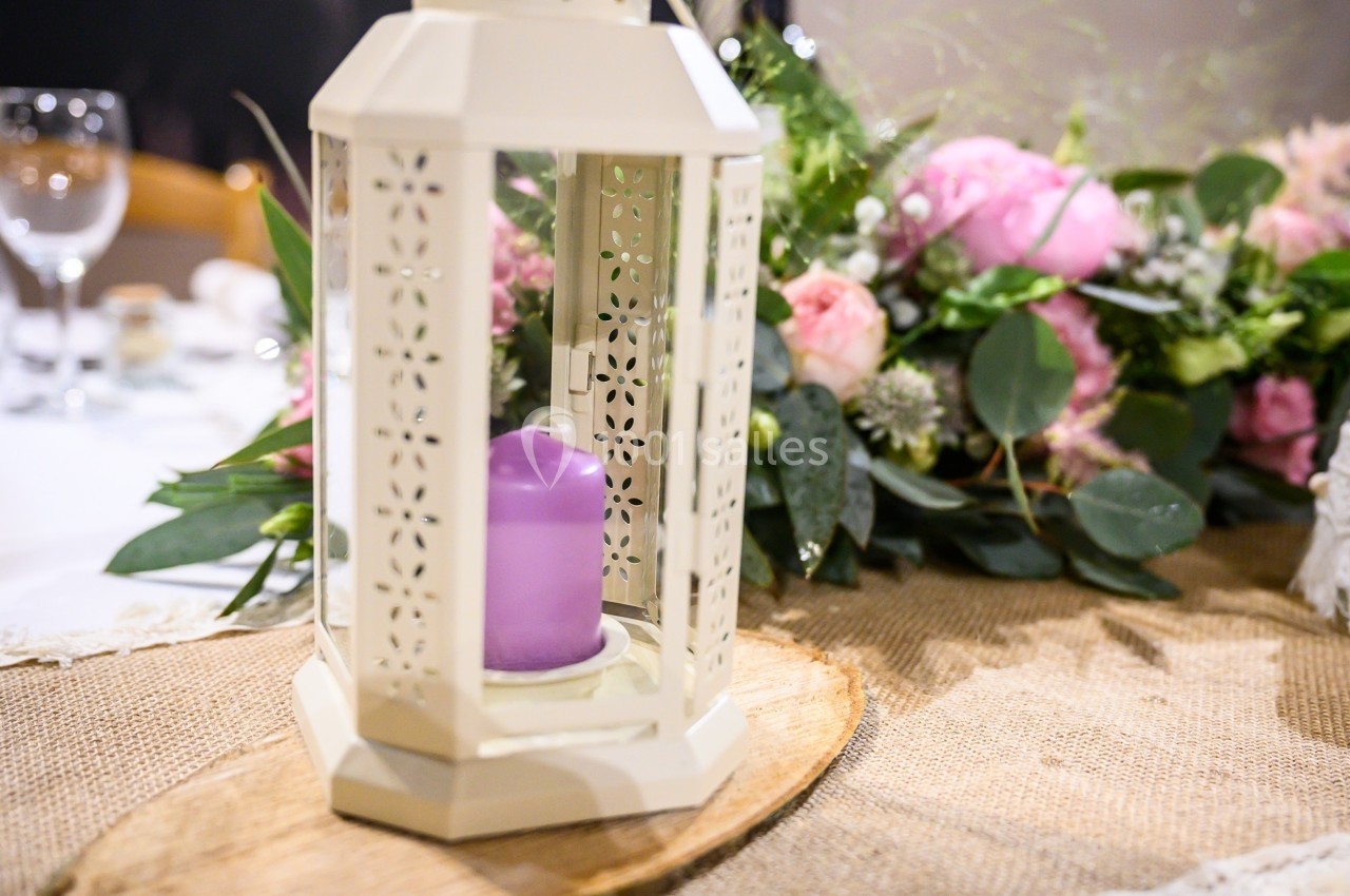 Lanterne blanche avec bougie violette sur une table décorée de fleurs et d'un chemin de table en toile de jute.