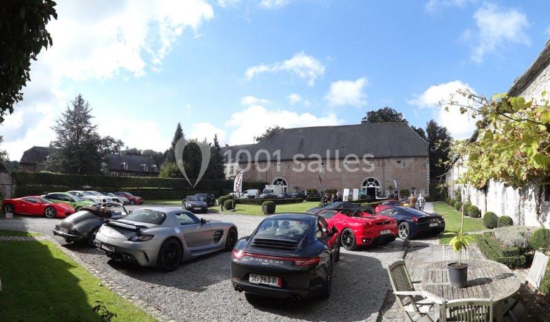 Plusieurs voitures de sport garées dans une cour pavée devant un bâtiment en briques avec un jardin environnant.
