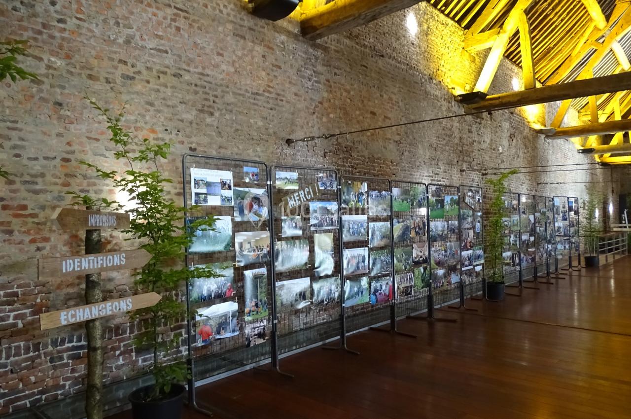 Exposition de photos accrochées sur des grilles dans une grande salle en briques avec un plafond en poutres apparentes.