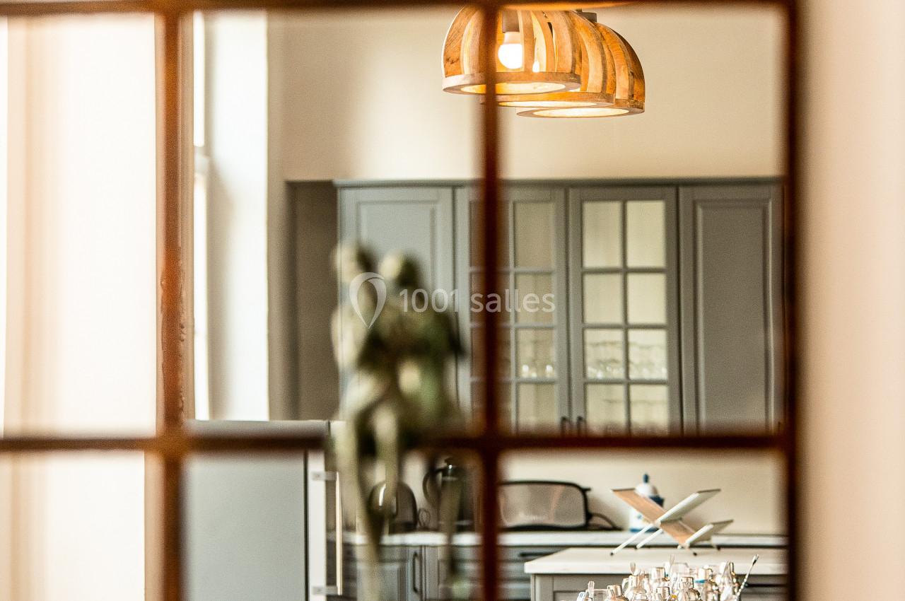 Vue d'une salle à manger élégante avec une table dressée, des chaises en osier et une cuisine en arrière-plan.