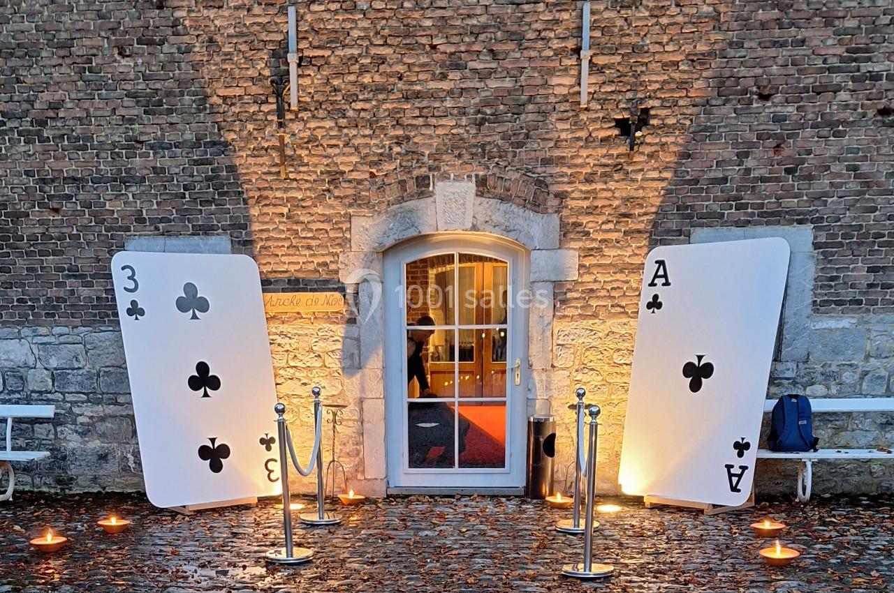 Deux grandes cartes à jouer (3 de trèfle et As de trèfle) encadrent une porte éclairée devant un mur en briques.