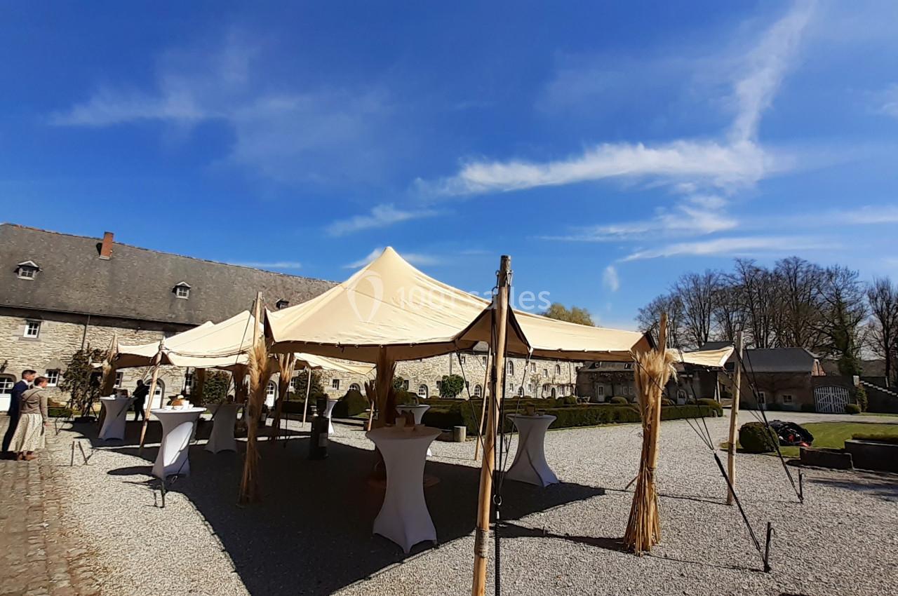 Tente beige installée dans une cour pavée, entourée de tables hautes et de bâtiments en pierre sous un ciel bleu.