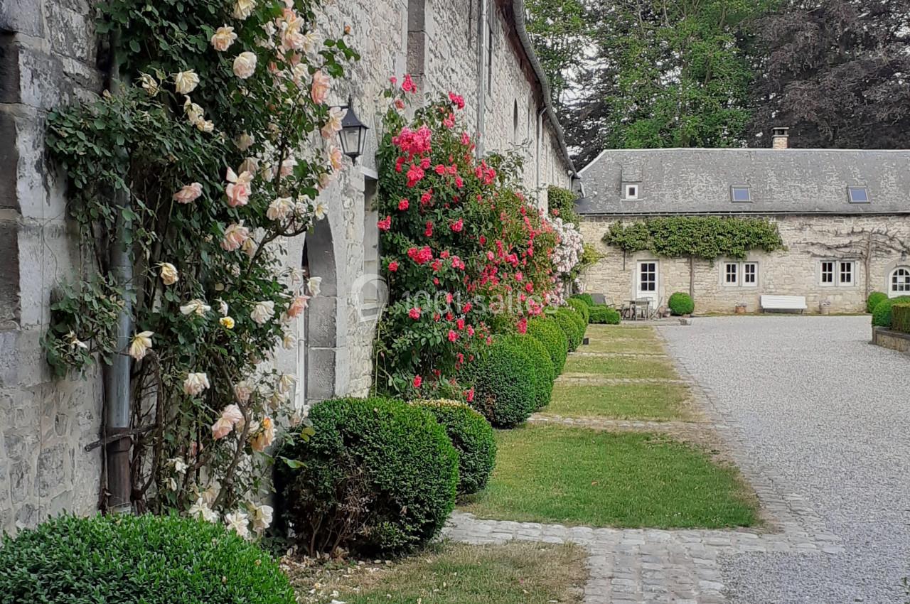 Façade en pierre d'un bâtiment ancien avec des rosiers grimpants, allée pavée et jardin soigné.