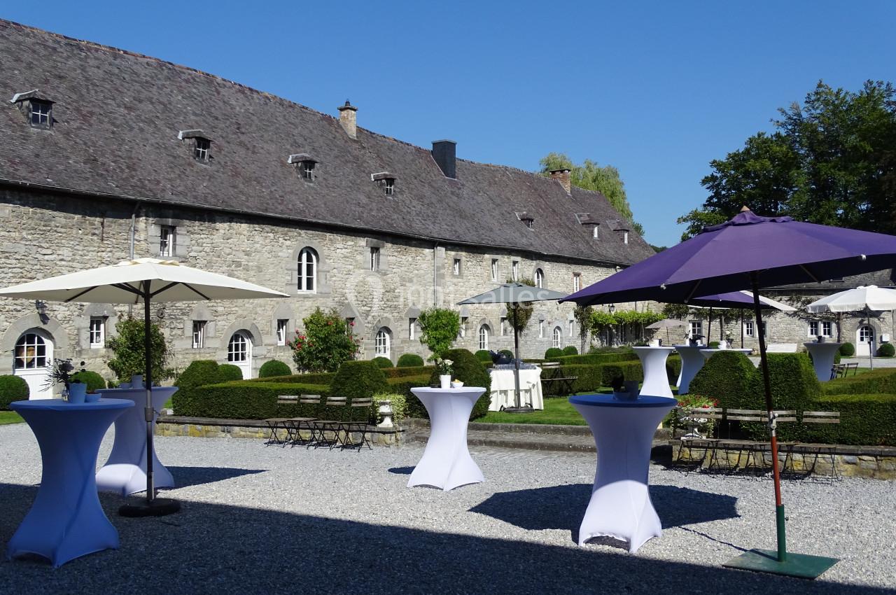 Cour d’un bâtiment en pierre avec tables hautes dressées et parasols colorés, entourée de haies et d’arbres.