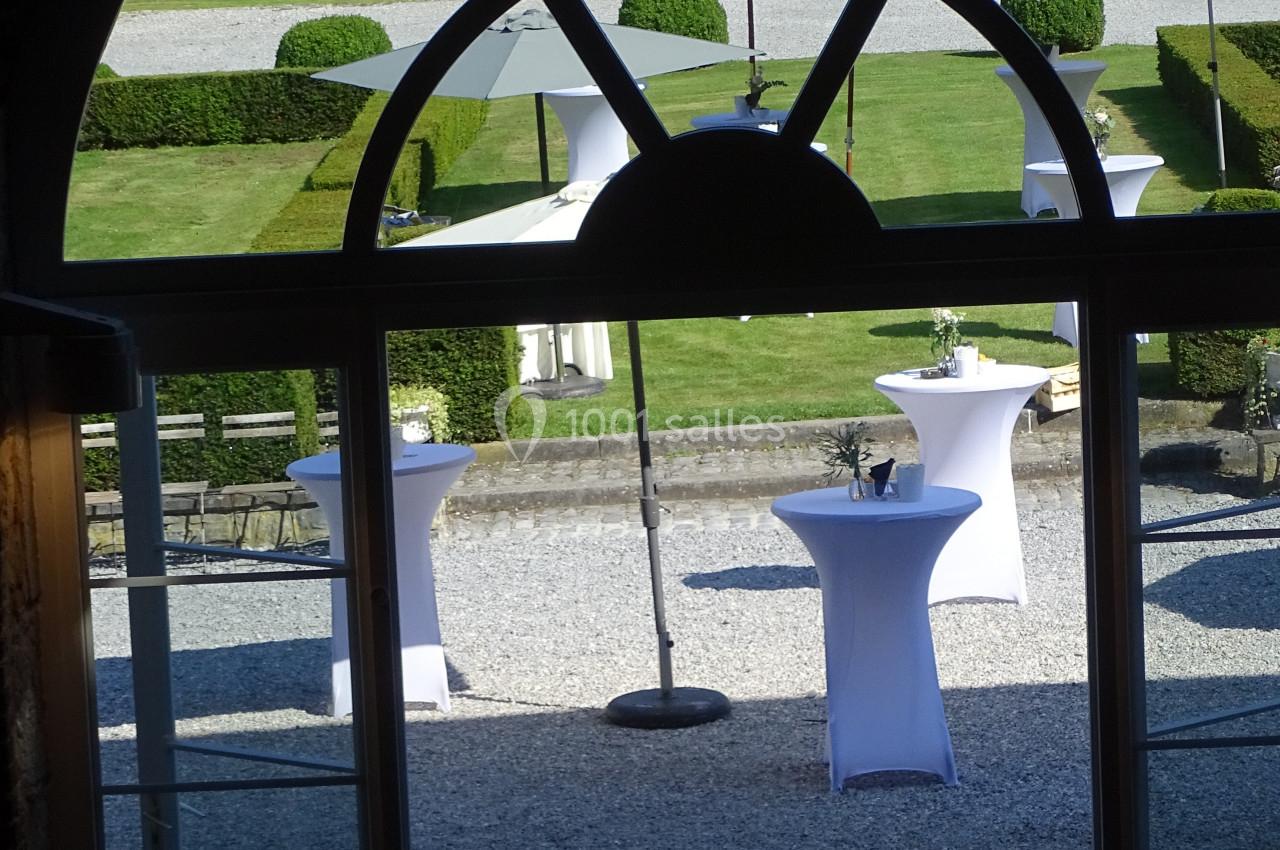 Vue depuis une porte vitrée sur une cour pavée avec des tables hautes dressées et un jardin en arrière-plan.