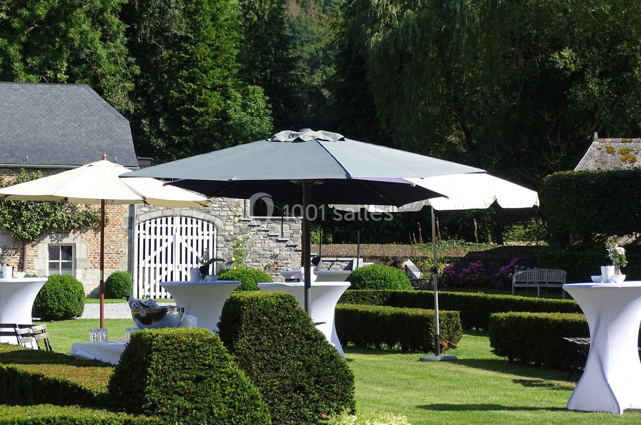 Parasols blancs et gris installés dans un jardin verdoyant avec des tables hautes et des haies taillées.