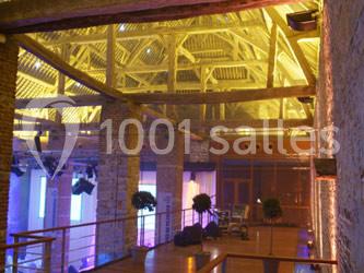 Vue intérieure d'une salle avec charpente en bois éclairée, murs en pierre et espace aménagé pour des événements.