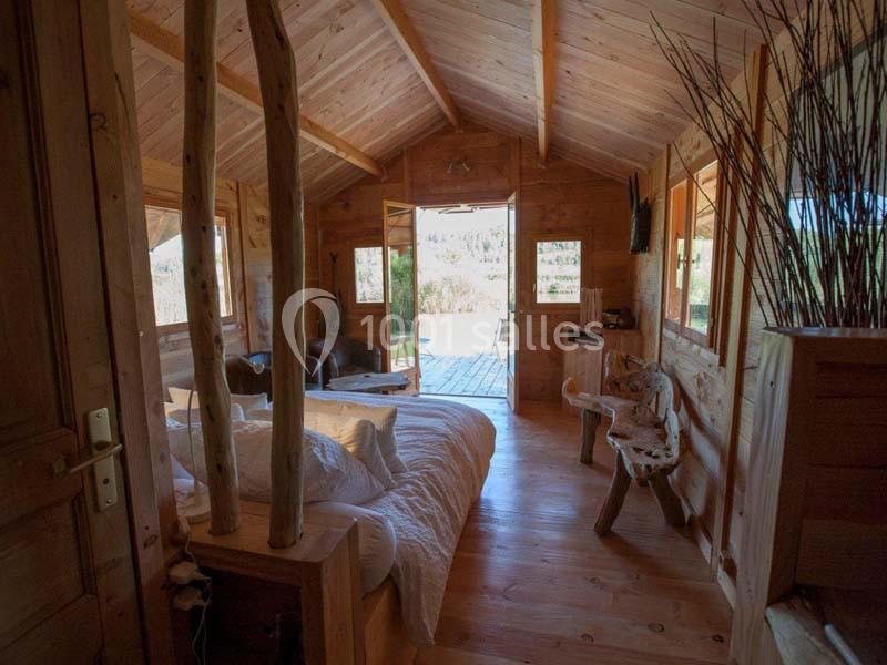 Intérieur d'un chalet en bois avec lit, mobilier rustique et vue sur une terrasse donnant sur un paysage naturel.