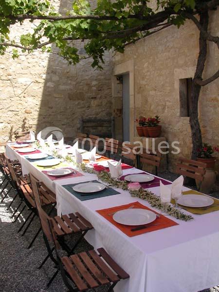 Table extérieure dressée avec nappes blanches, assiettes et serviettes, sous une pergola dans une cour en pierre.