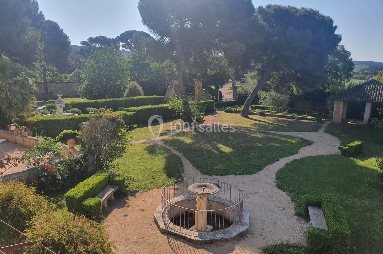 Jardin paysager avec allées en gravier, fontaine centrale protégée par une grille et arbres en arrière-plan.