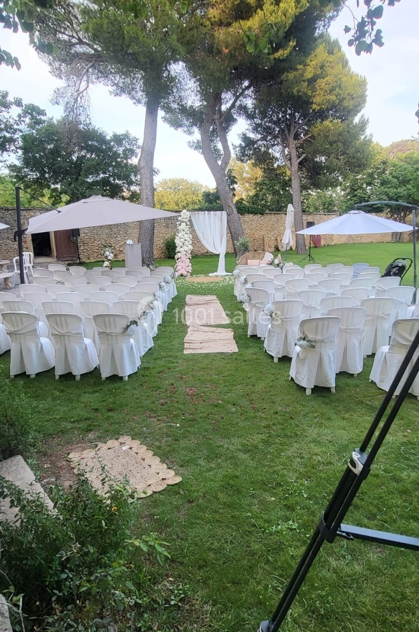 Chaises blanches alignées en extérieur pour une cérémonie, avec une allée centrale et un décor floral sous des arbres.