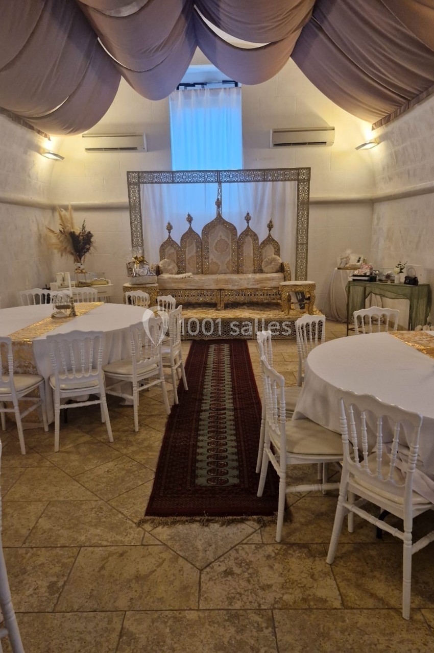 Salle décorée pour un événement avec tables rondes, chaises blanches et une estrade ornée de motifs orientaux.