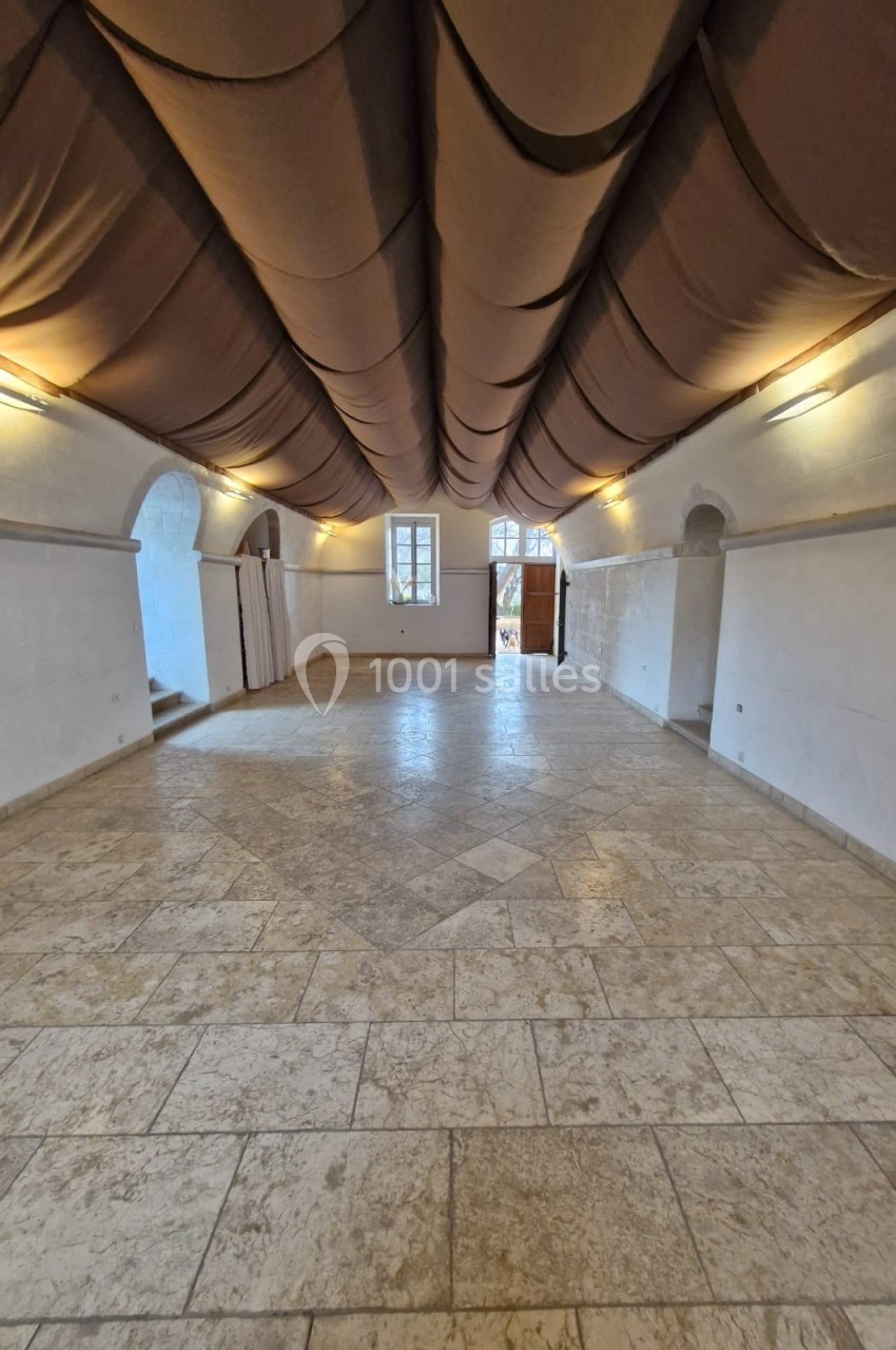 Salle vide avec un sol en pierre, plafond voûté recouvert de tissu beige et murs blancs, éclairée par des fenêtres et des…