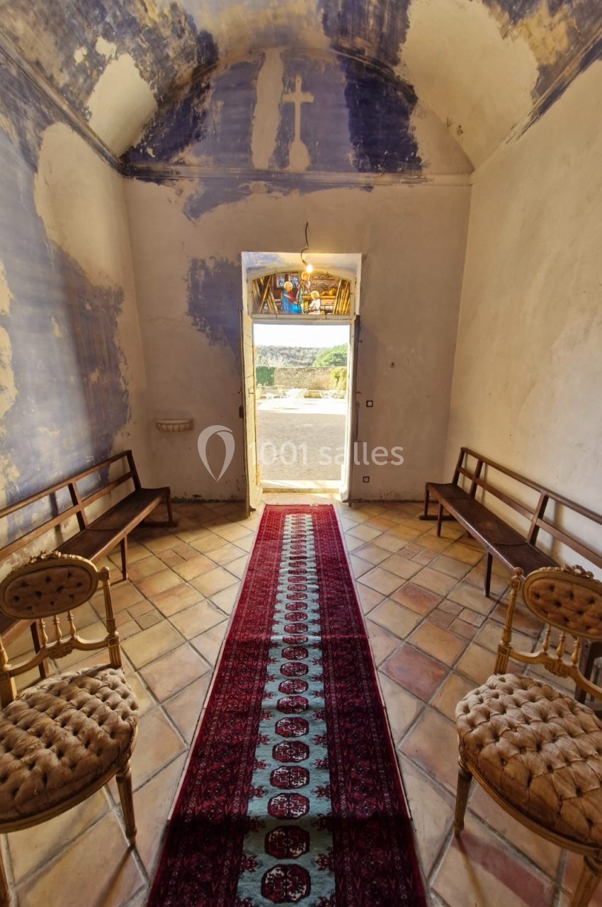 Intérieur d'une petite chapelle avec un tapis rouge central, des bancs en bois et une porte ouverte sur un paysage lumineux.