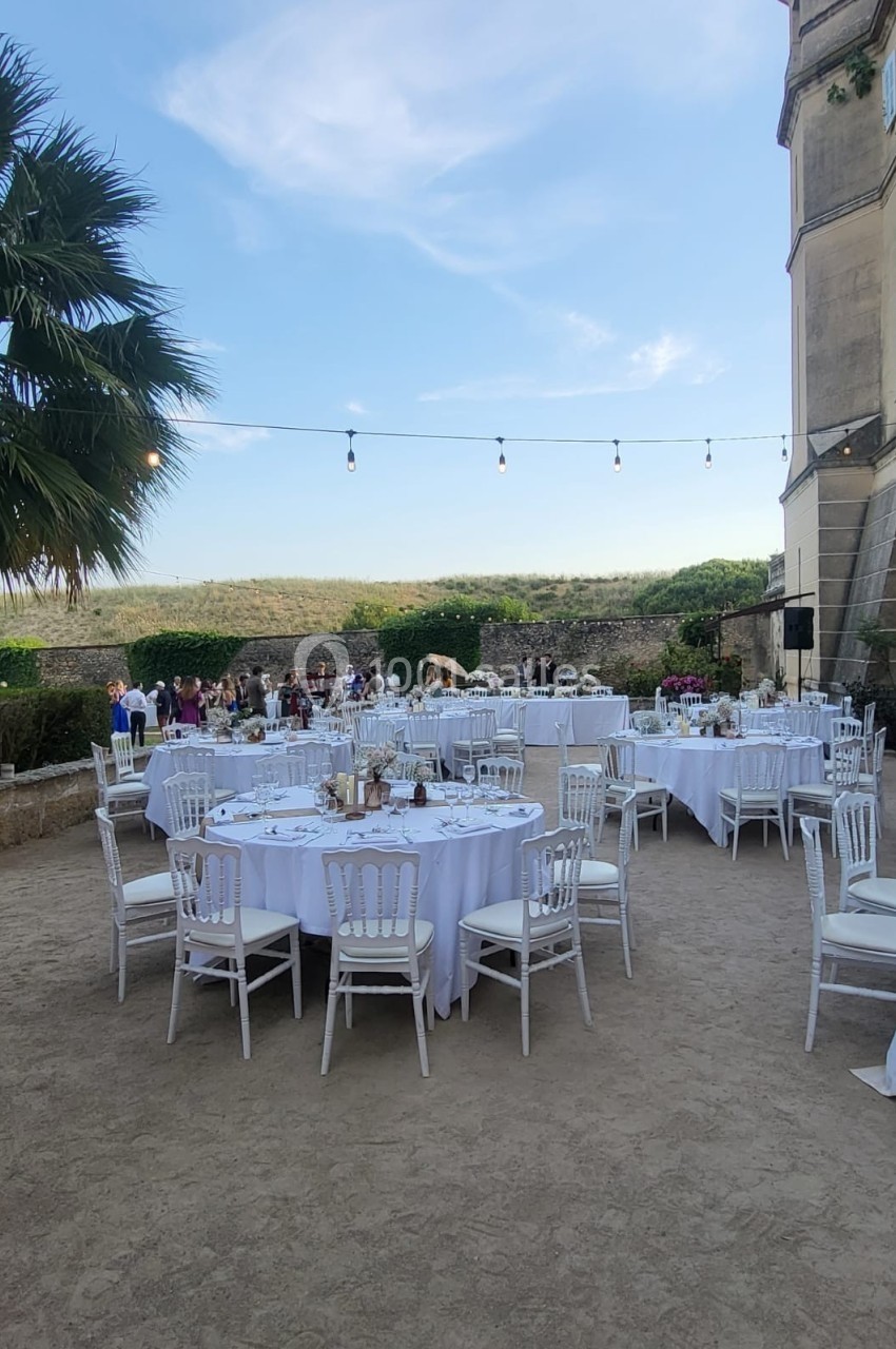 Tables rondes dressées avec nappes blanches dans une cour extérieure, près d'un bâtiment ancien, sous un ciel dégagé.