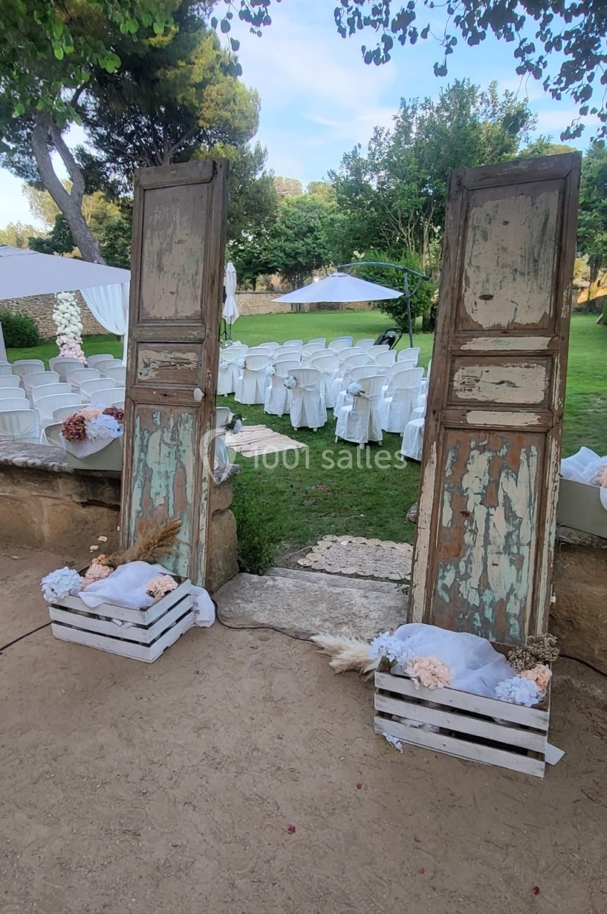 Deux portes en bois vieilli encadrent une allée menant à des chaises blanches disposées pour une cérémonie en extérieur.