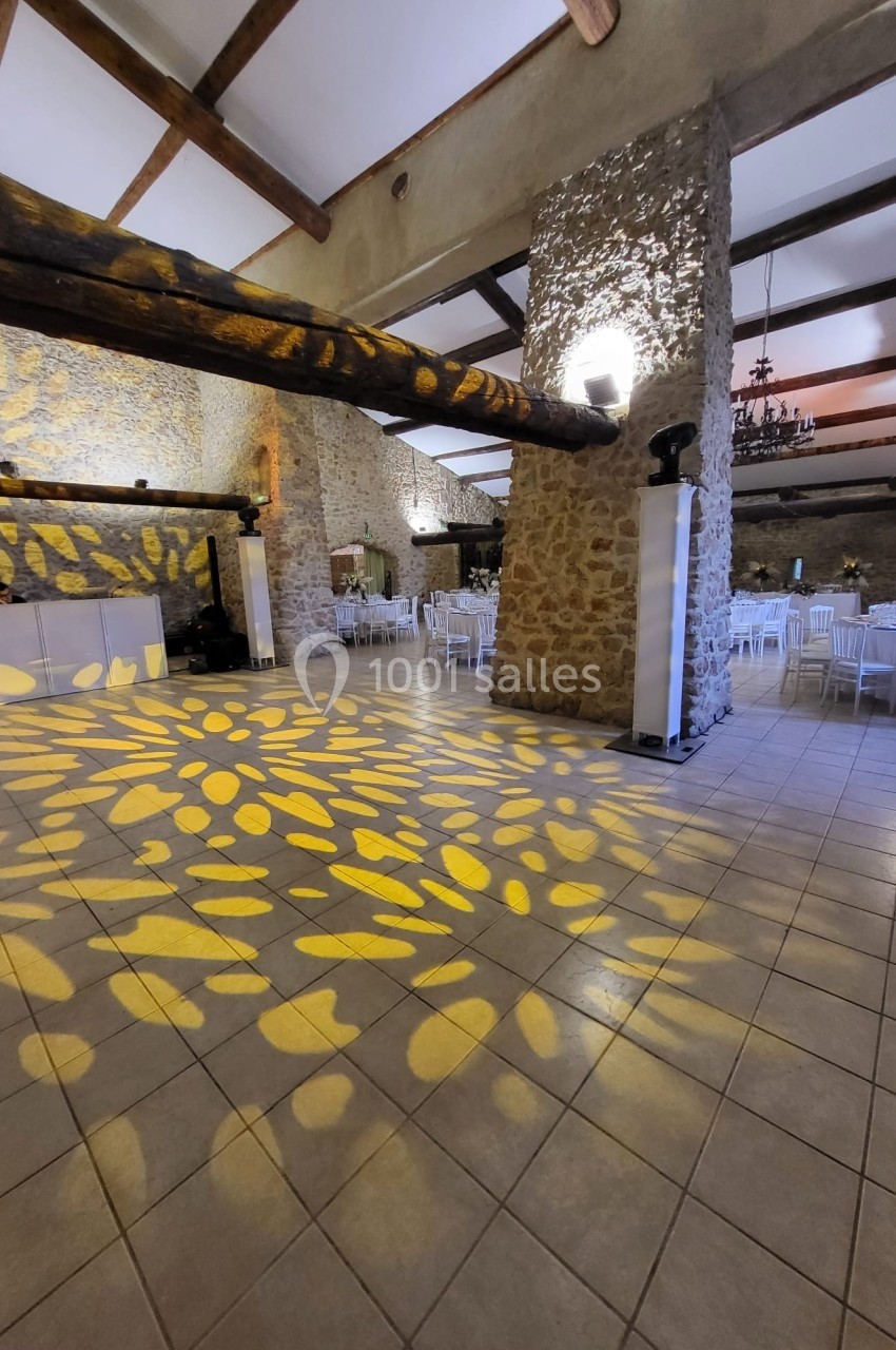 Salle en pierre avec poutres apparentes, éclairage décoratif au sol et chaises blanches disposées pour un événement.