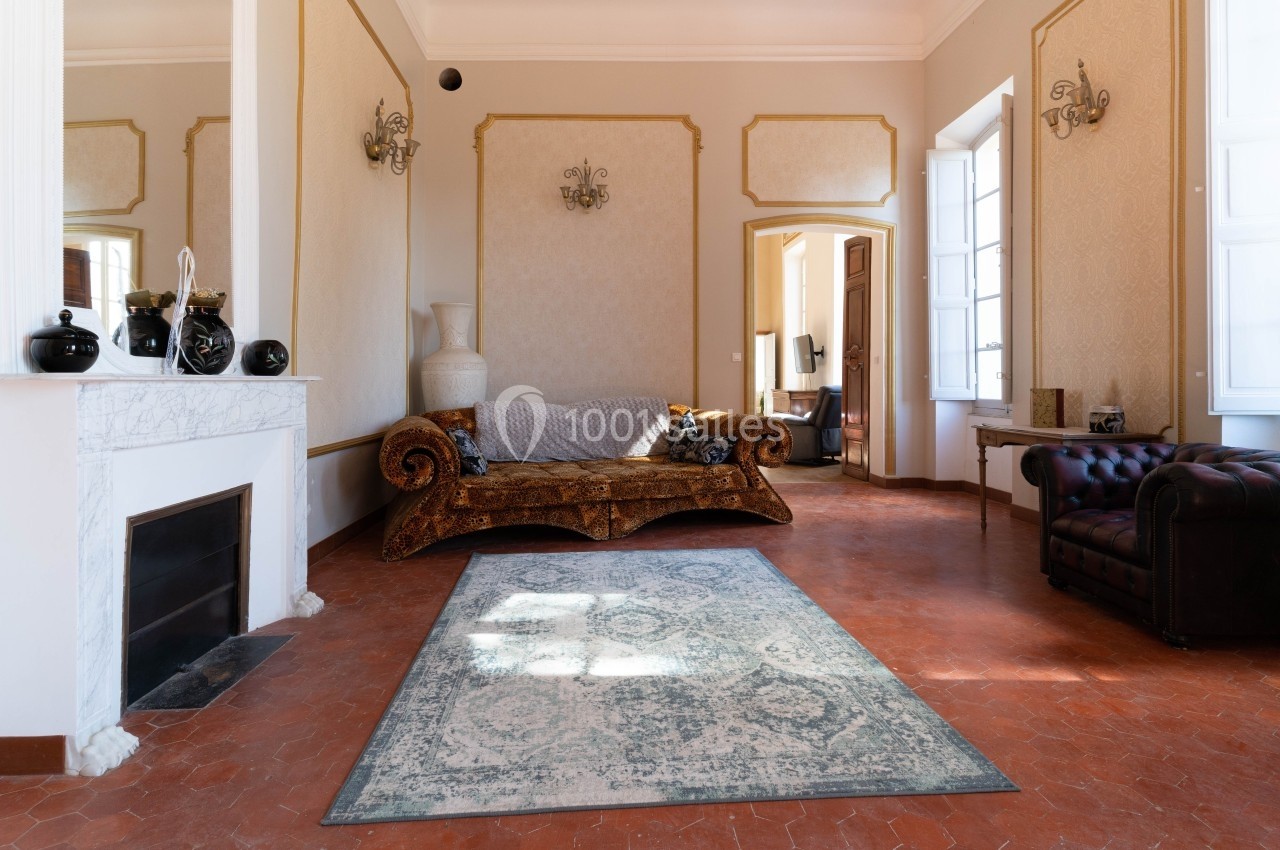 Salon lumineux avec canapé en velours, cheminée, tapis bleu et grandes fenêtres laissant entrer la lumière naturelle.