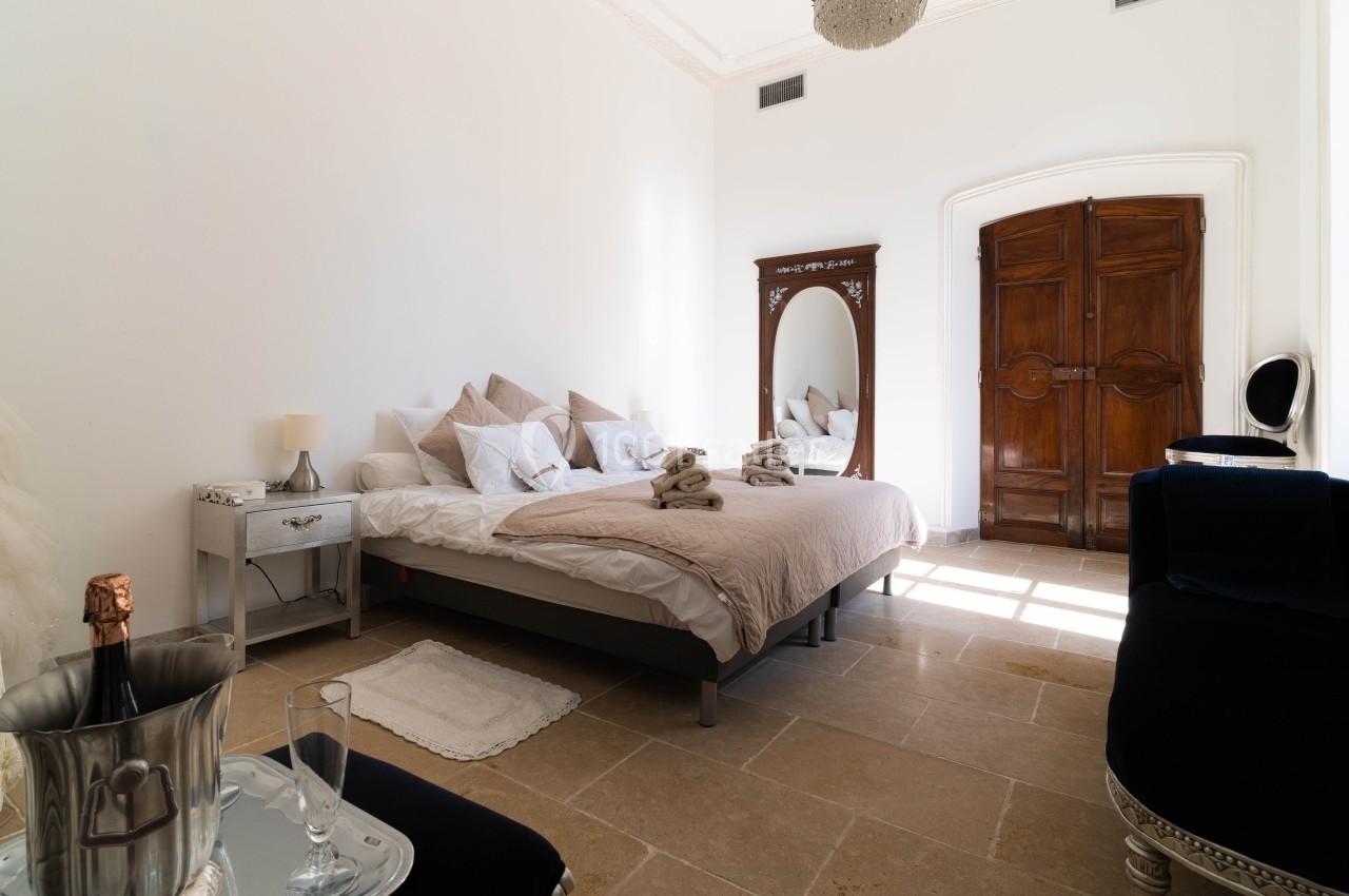 Chambre lumineuse avec lit double, miroir ancien, porte en bois, sol en pierre et seau à champagne sur une table.