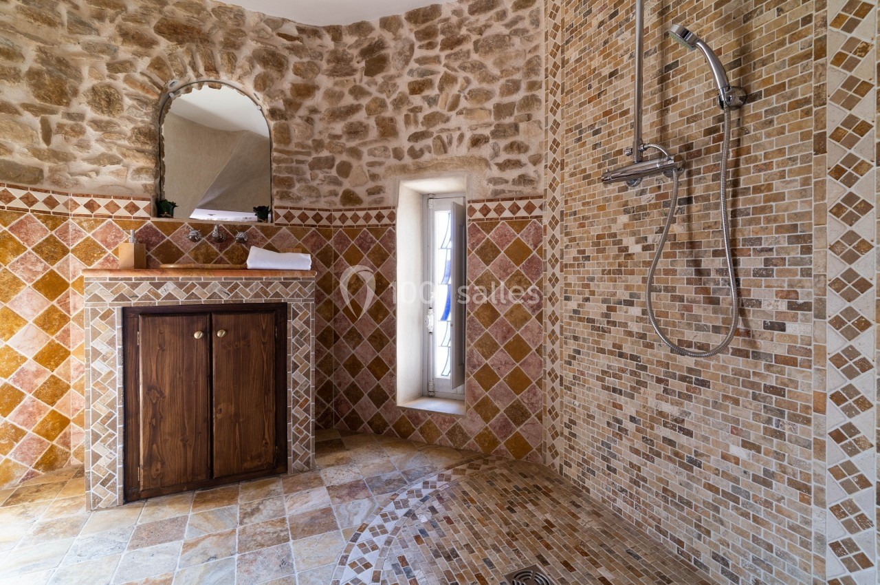 Salle de bain avec murs en pierre et mosaïque, douche à l'italienne et meuble en bois sous un miroir.