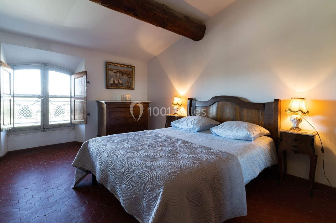 Chambre avec lit double, couvre-lit blanc, meubles en bois, lampes allumées et fenêtre laissant entrer la lumière naturelle.