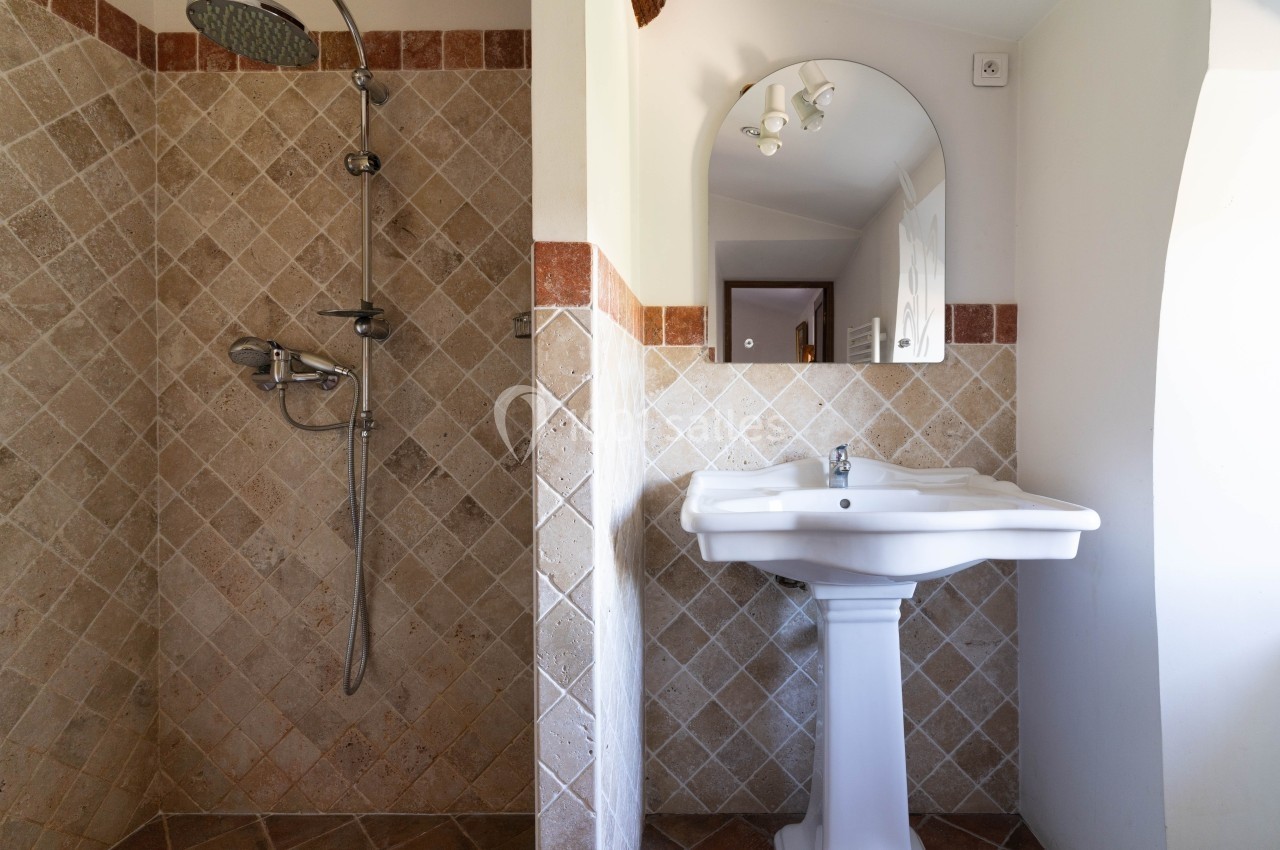 Douche avec carrelage beige et lavabo blanc sur pied, surmonté d'un miroir dans une salle de bain lumineuse.