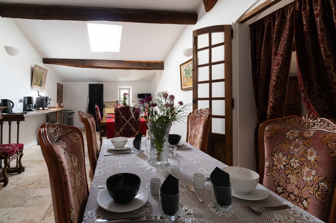 Salle à manger lumineuse avec table dressée, chaises ornées, poutres apparentes et vue sur une cuisine équipée.