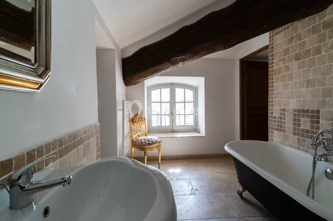 Salle de bain lumineuse avec baignoire sur pieds, lavabo moderne, mur en carreaux beige et poutre apparente.