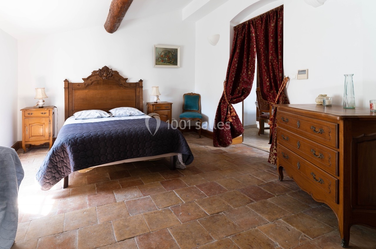 Chambre lumineuse avec lit double, meubles en bois ancien, sol en terre cuite et rideaux rouges.