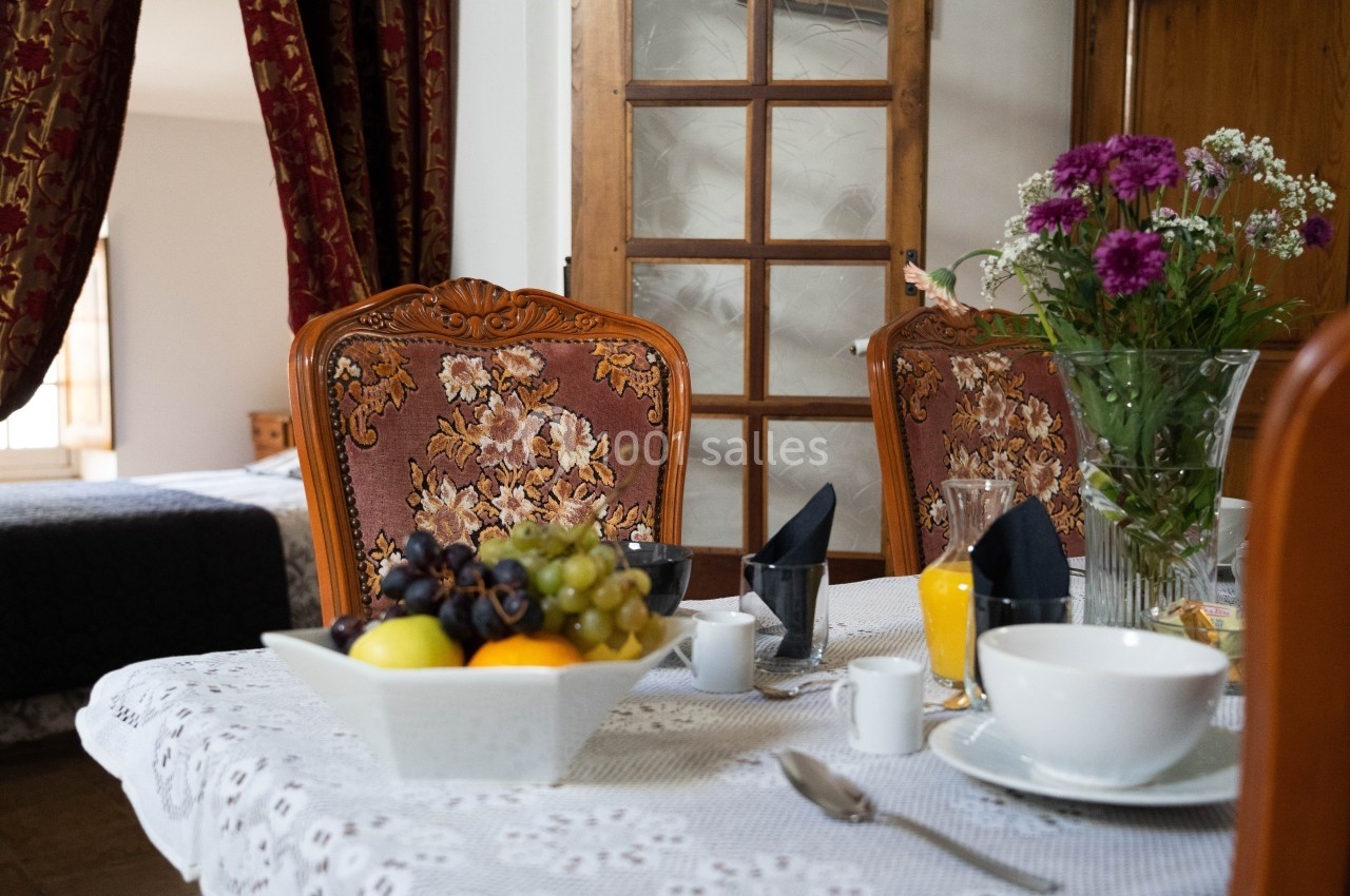 Table dressée avec une nappe blanche, contenant un bol de fruits, des fleurs, des tasses et un verre de jus d'orange.