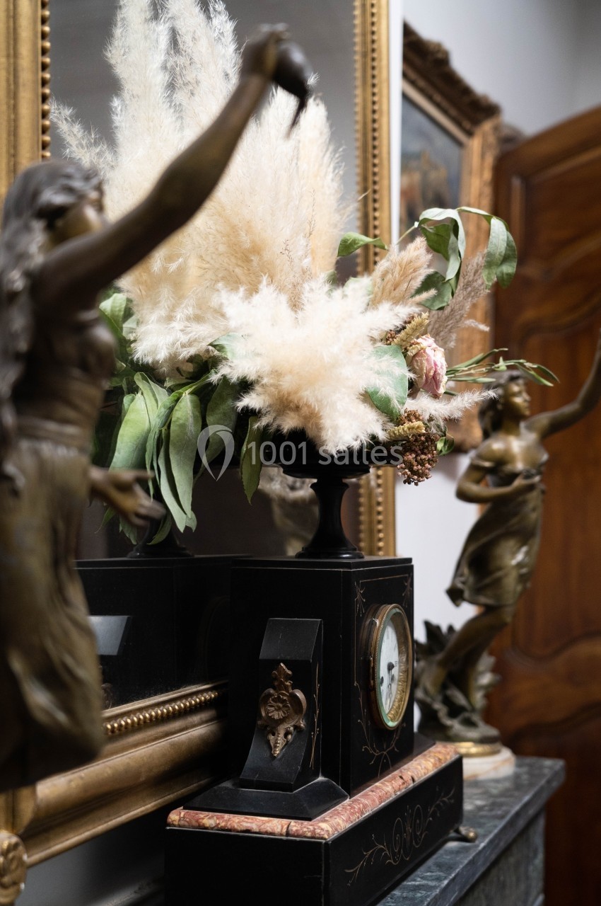 Composition florale avec des herbes de la pampa dans un vase noir, entourée de statues en bronze et d'un cadre doré.